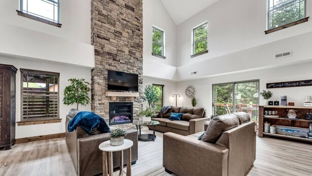 A spacious living room with a stone fireplace, comfortable seating, and large windows offering views of the outdoors. - Creekside Cabin / Branson, SDC, Lakes, In/Out Pool (Hollister)