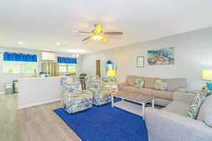 A living room with a sofa, two armchairs, a coffee table, a ceiling fan, and a kitchen area in the background. - Tilghman Ranch Retreat (North Myrtle Beach)