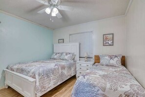 A bedroom with two beds, a ceiling fan, a window with blinds, and a nightstand with a lamp.