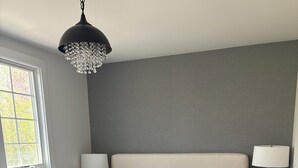 Ein ordentlich bezogenes Bett mit weißen Leinen und einem gefalteten Handtuch, ein Nachttisch mit Lampe, eine graue Wand und ein Fenster mit Tageslicht.