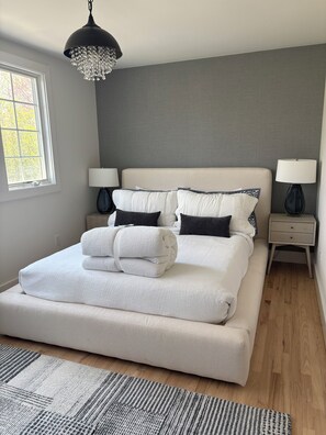 A neatly made bed with white linens and a folded towel, a bedside table with a lamp, a grey wall, and a window with daylight.