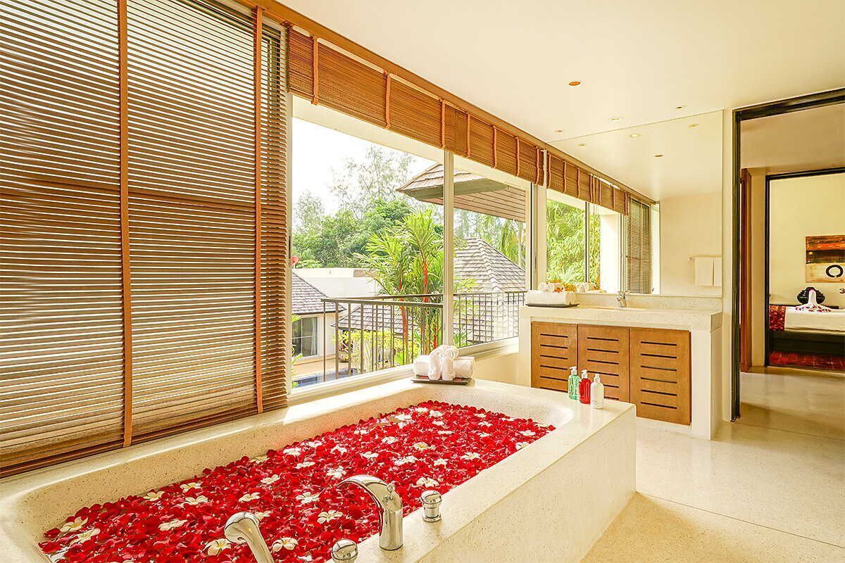 A modern bathroom with a large bathtub filled with red rose petals, wooden cabinets, and a view of greenery outside.