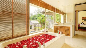 A modern bathroom with a large bathtub filled with red rose petals, wooden cabinets, and a view of greenery outside. - Villa Samakee (Choeng Thale)