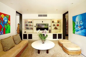 A living room with a sofa, ottoman, TV, bookshelf, and decorative items. - Villa Samakee (Choeng Thale)