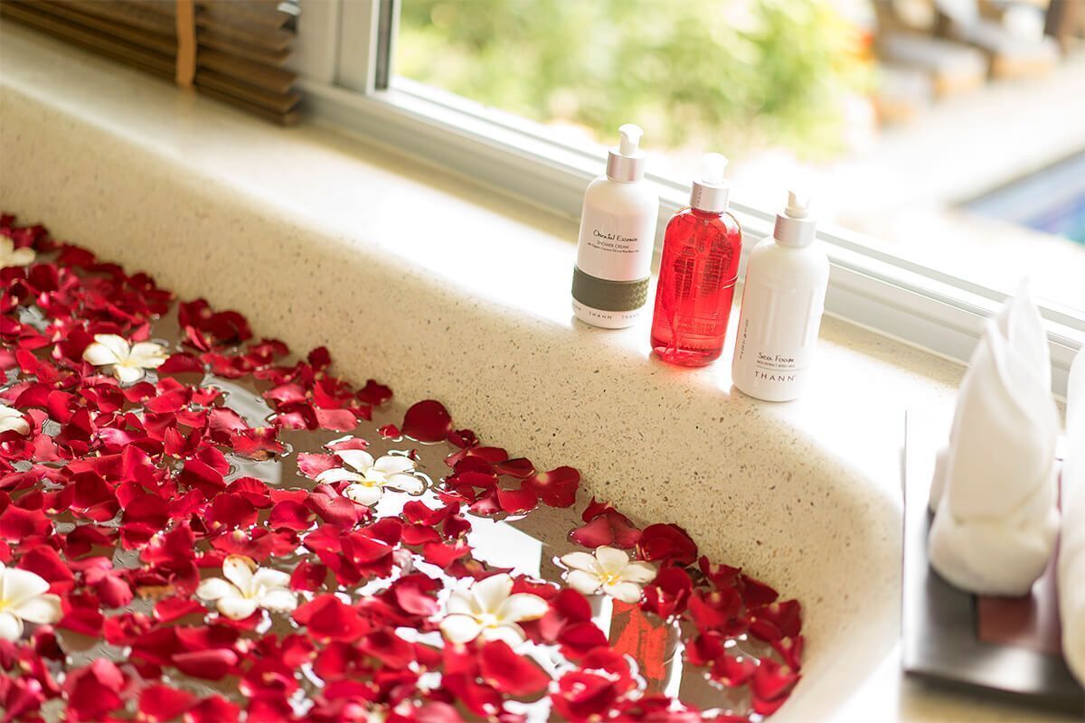 A bathtub filled with rose petals and two bottles of bath products on a windowsill.
