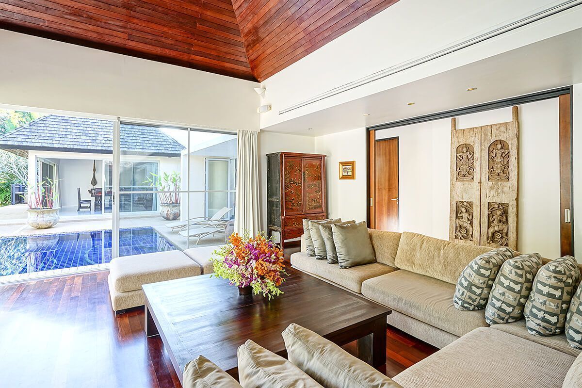 A spacious living area with a wooden ceiling, a large sofa set, a coffee table, and a view of a swimming pool.