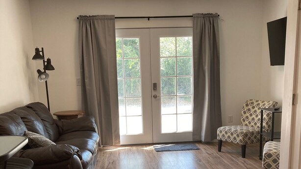 A spacious living room with a leather sofa, a small table, and a patterned armchair. A large window with curtains allows natural light to enter. - Goodturn Cottage (Cody)
