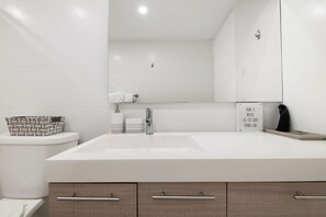 A modern bathroom with a large white sink, a mirror above it, and a toilet with a patterned basket on top. There are two towels hanging on the wall and a sign with a quote. - Coastal Living With Golf Course Vistas Otium (Humacao)