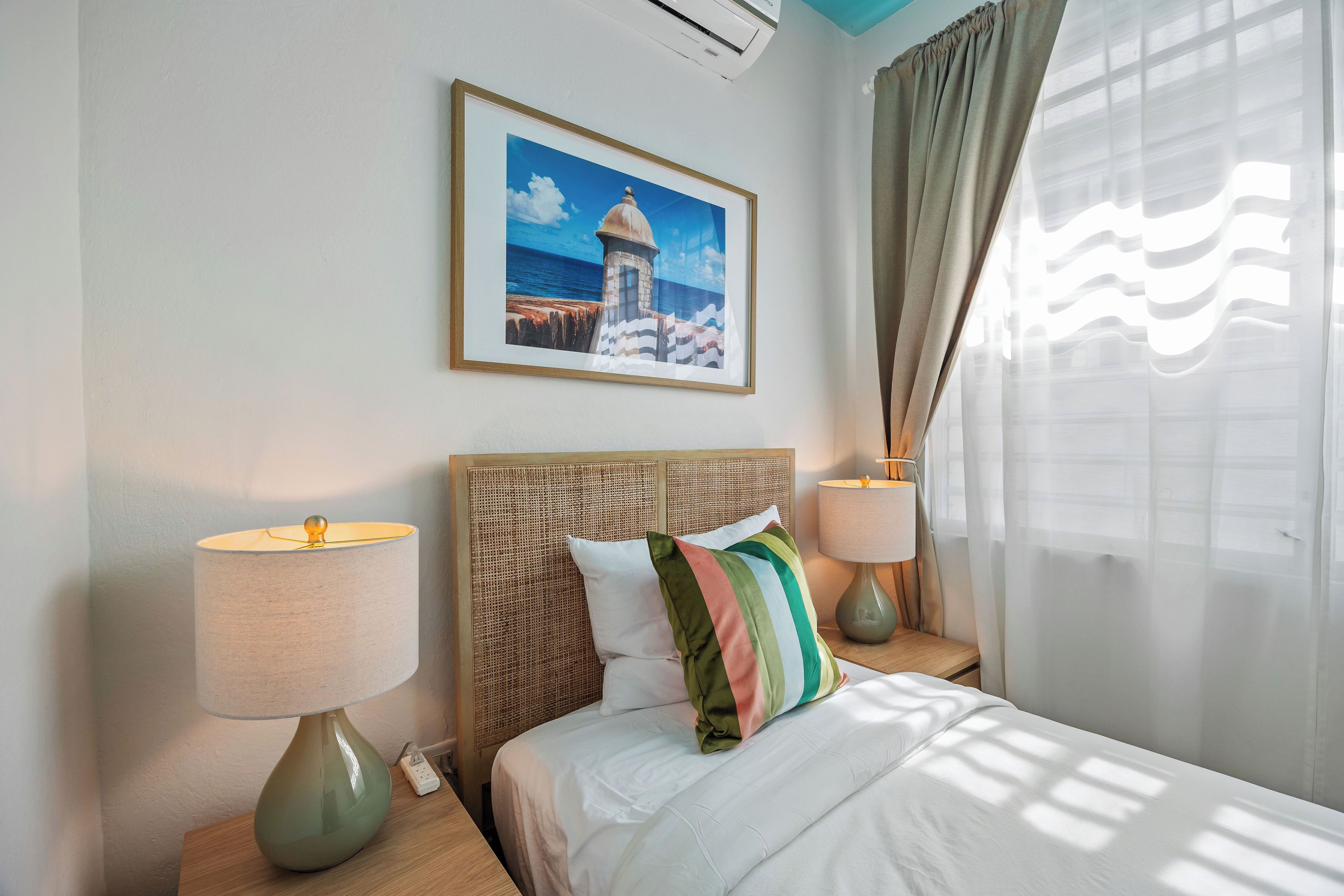A hotel room with a bed, two bedside lamps, a framed picture on the wall, and sheer curtains.