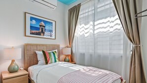 A neatly arranged bedroom with a bed, bedside tables, a lamp, a framed picture, and sheer curtains. - Luxury Condado Apt Walk to the Beach Pool (San Juan)