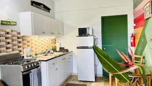 A compact kitchen with white cabinets, a stove, a refrigerator, and a microwave. There is a green door and a plant in the corner.