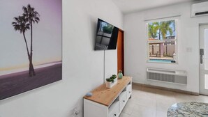 A modern living room with a flat-screen TV mounted on the wall, a wooden console table with decorative items, a large window with a view of palm trees and a pool, and a tiled floor. - Coastal Home Safe Secure Parking (San Juan)