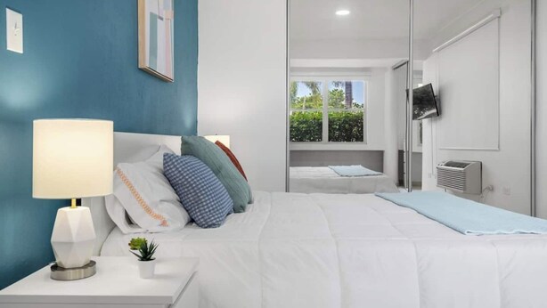 A modern bedroom with a bed, bedside table, lamp, and a view of greenery through a window. - Coastal Home Safe Secure Parking (San Juan)
