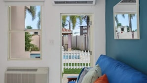 A room with a blue couch, a white air conditioner, a window with a view of a pool and palm trees, and a white fence. - Coastal Home Safe Secure Parking (San Juan)