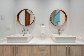 Un baño con dos lavamanos, cada uno con un espejo redondo encima. Debajo de los lavamanos hay armarios de madera, y se ve un dispensador de toallas entre los espejos.