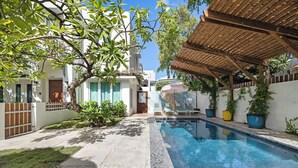 A swimming pool with a wooden pergola, surrounded by a white building with a green lawn and potted plants. - In Condado Beach Beautiful Pool With Tanning Bed (San Juan)