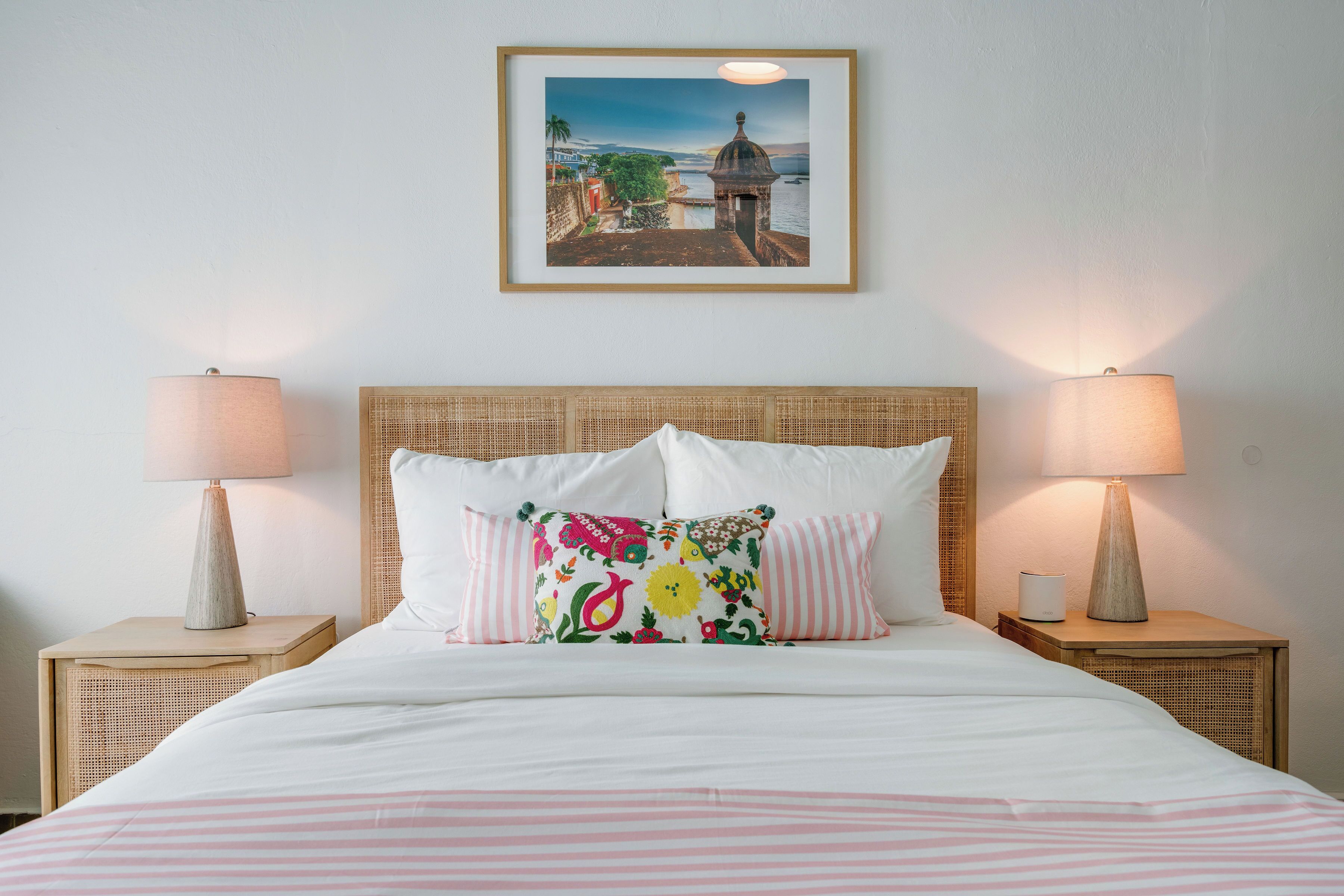 A neatly made bed with white bedding and pink stripes, flanked by two bedside tables with lamps, a wicker headboard, and a framed picture of a coastal scene above the bed.