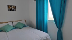 Un lit avec une literie blanche et des coussins turquoise, une tête de lit en bois et une fenêtre avec des rideaux bleus.