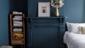 A bedroom with a dark blue wall, a wooden shelf with folded towels, a framed picture above a fireplace, and a vase with flowers on a side table. - Group-Friendly 4BR / 3.5BA / 2 Kitchens + Private Homes Side-by-Side  Sleeps 10 (Dayton)