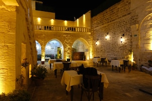 Un área de comedor al aire libre con muros y arcos de piedra, mesas con manteles amarillos y plantas en macetas.
