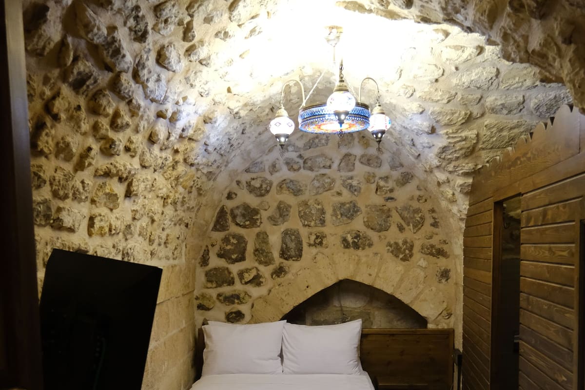 A stone-walled room with a bed, a wooden headboard, a patterned rug, and a ceiling light fixture.