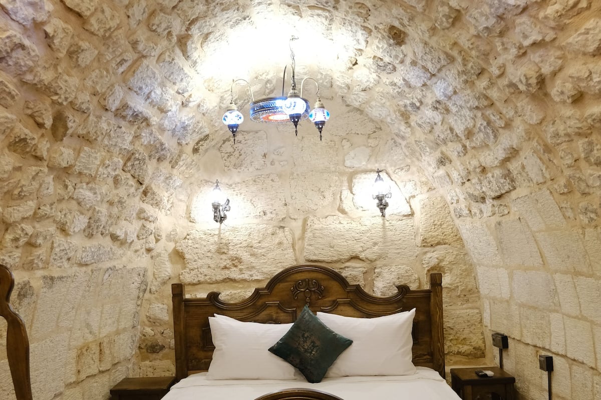 A stone-walled bedroom with a wooden bed, a patterned rug, and wall-mounted lamps.