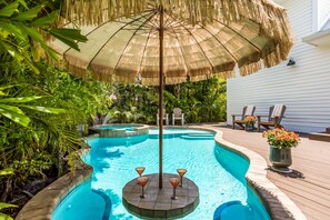 A swimming pool with a thatched umbrella, surrounded by lush greenery and deck chairs. - Beach Dream Cottage by Beach Retreats (Bradenton Beach)