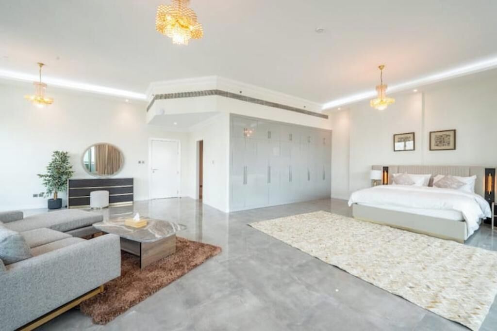 Ein modernes Schlafzimmer mit einem großen Bett, einer Couch, einem Couchtisch und einem Spiegel. Ein Kleiderschrank mit Spiegel darüber, und eine Tür zu einem weiteren Raum.
