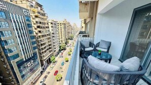 Uma varanda com mesa de vidro e duas cadeiras, com vista para uma rua com carros estacionados e edifícios identificados com "G2".