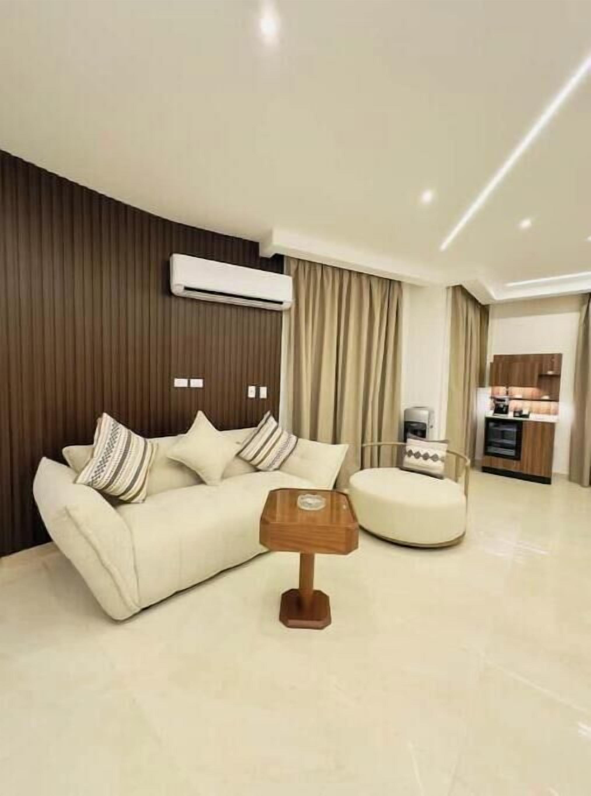 A modern living room with a sofa, ottoman, and a wooden side table. There is a kitchen area in the background.