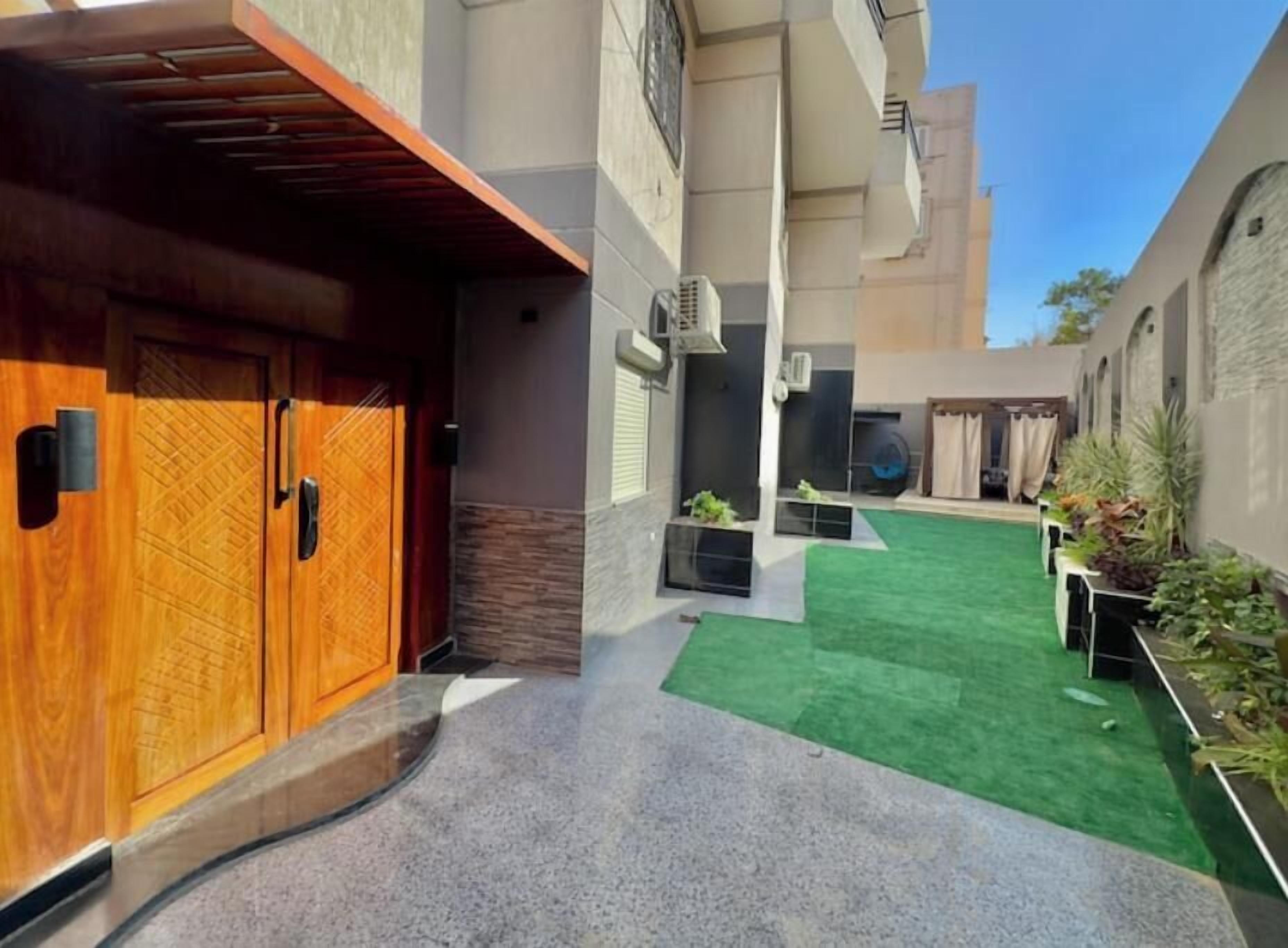 Une cour avec une porte en bois, une pelouse artificielle verte et un petit espace jardin avec des plantes.
