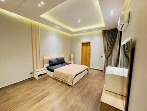 Une chambre à coucher moderne avec un plancher en bois, un lit recouvert d’une couvre-lit beige et noire, une table de chevet, une armoire et un luminaire au plafond.