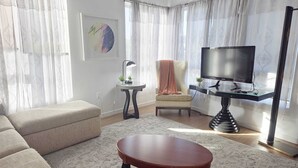 Una sala de estar con un sofá, una mesa de centro, un televisor sobre un mueble, una silla con una manta y una pequeña mesita de noche con una lámpara.