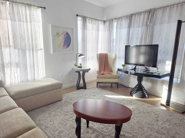 Ein Wohnzimmer mit einer Couch, einem Couchtisch, einem Fernseher auf einem Schrank, einem Sessel mit Decke und einem kleinen Beistelltisch mit Lampe.