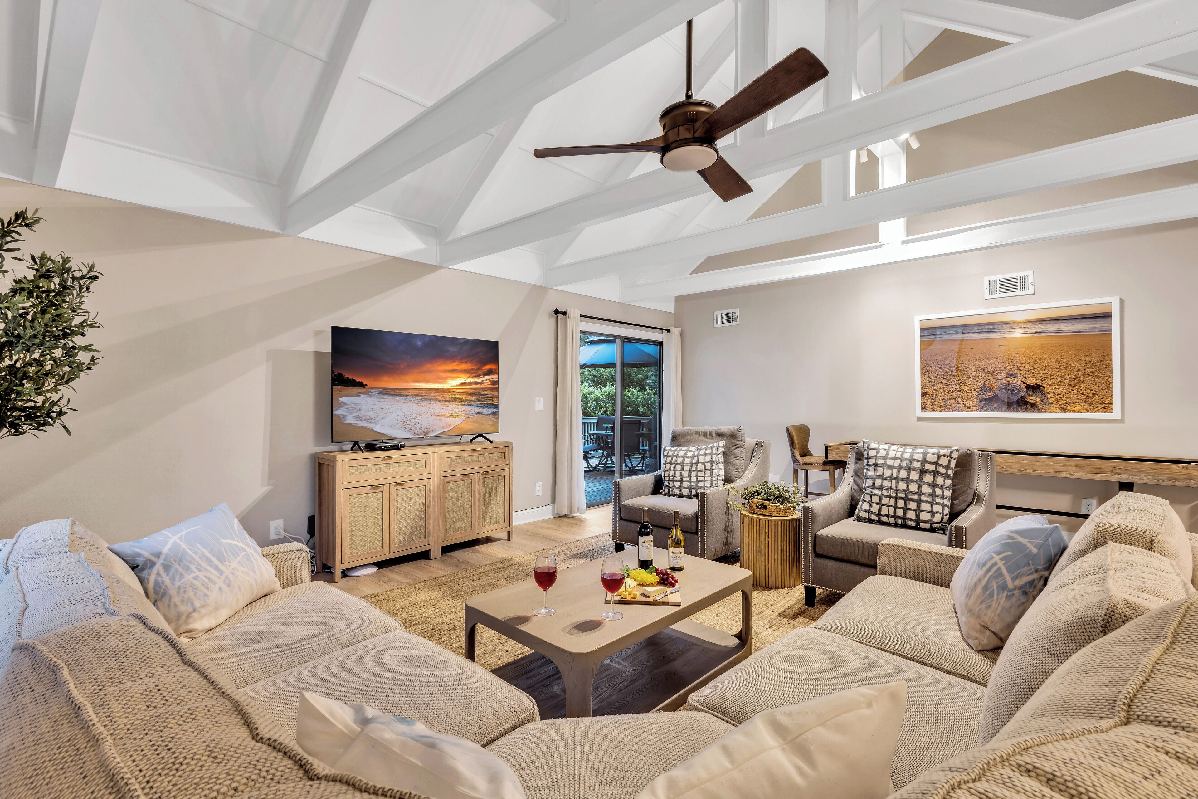 A spacious living room with a high ceiling, a ceiling fan, a large window with a view of the outdoors, a wooden cabinet, a sofa set, a coffee table with wine glasses and fruit, and a painting on the wall.