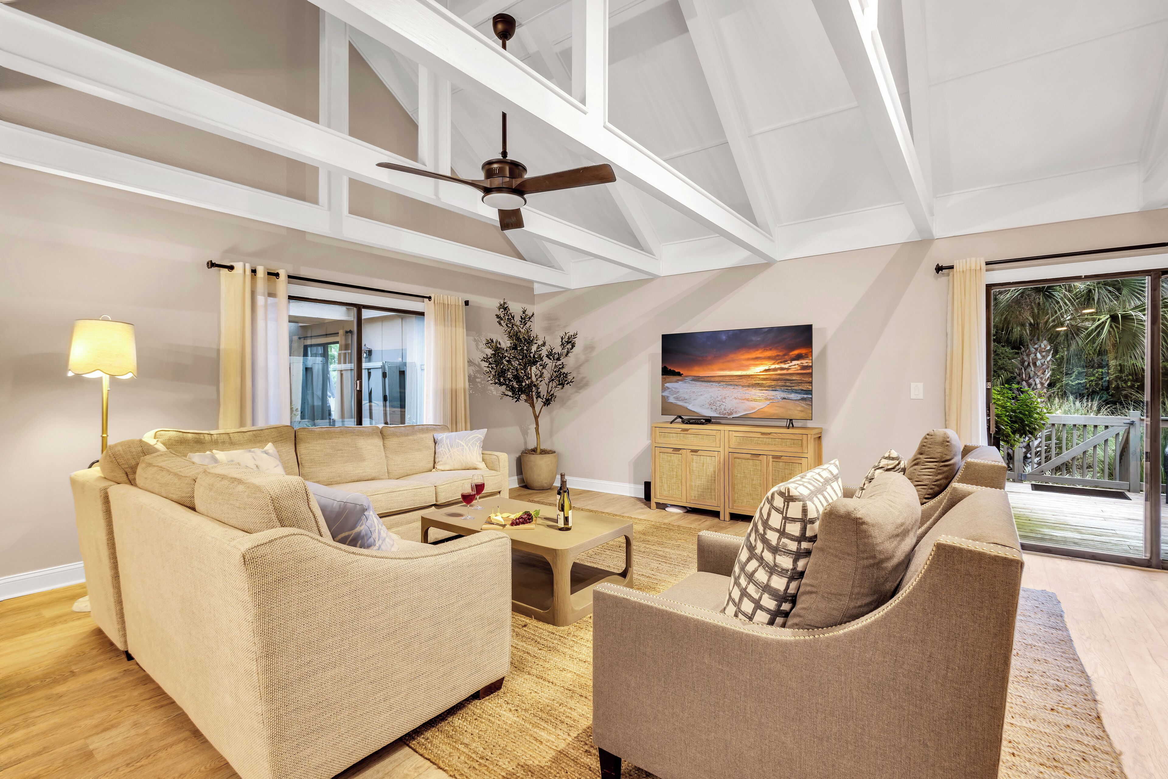 Ein geräumiges Wohnzimmer mit hoher Decke, Deckenventilator, großem Fenster mit Vorhängen, einem beigefarbenen Sofa, einem Couchtisch und einem Gemälde, das einen Sonnenuntergang zeigt.