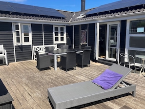 A covered outdoor area with a table and chairs, a lounge chair, and a sliding glass door.