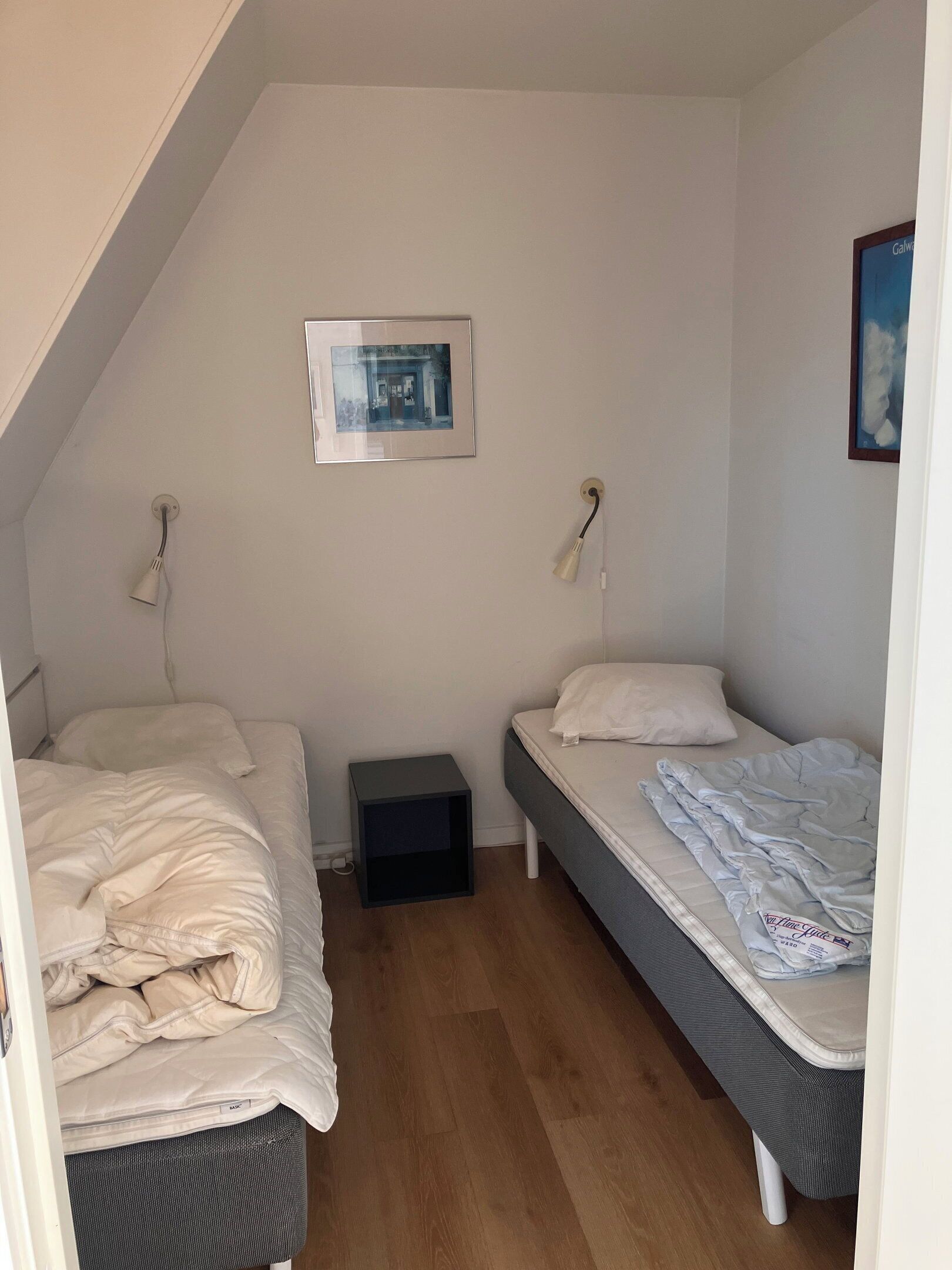 Ein kleines, schlichtes Zimmer mit zwei Betten, einem kleinen Tisch und einem gerahmten Bild an der Wand.