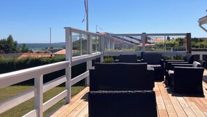 Una terraza con asientos al aire libre, barandilla y vista al mar y edificios.