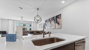 Une cuisine moderne dotée d’un grand îlot central, d’un évier en acier inoxydable et d’un lave-vaisselle. Des œuvres d’art encadrées ornent le mur, et un luminaire est fixé au plafond.