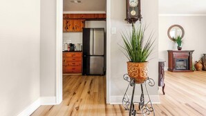 Un pasillo con piso de madera que conduce a una cocina con armarios de madera y electrodomésticos de acero inoxidable. Hay una planta en maceta sobre un soporte de metal en el pasillo.