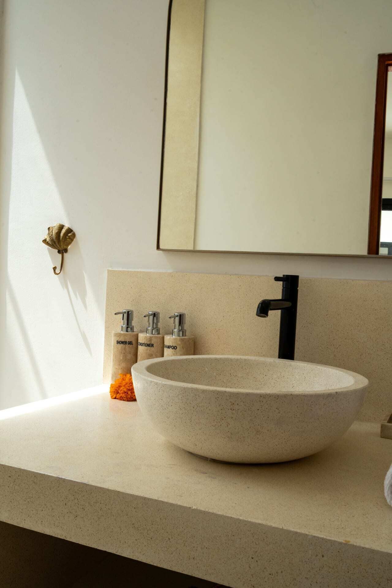 A modern bathroom with a large, round, beige sink, a black faucet, and three bottles of toiletries labeled "shower gel," "conditioner," and "shampoo" on a beige countertop.