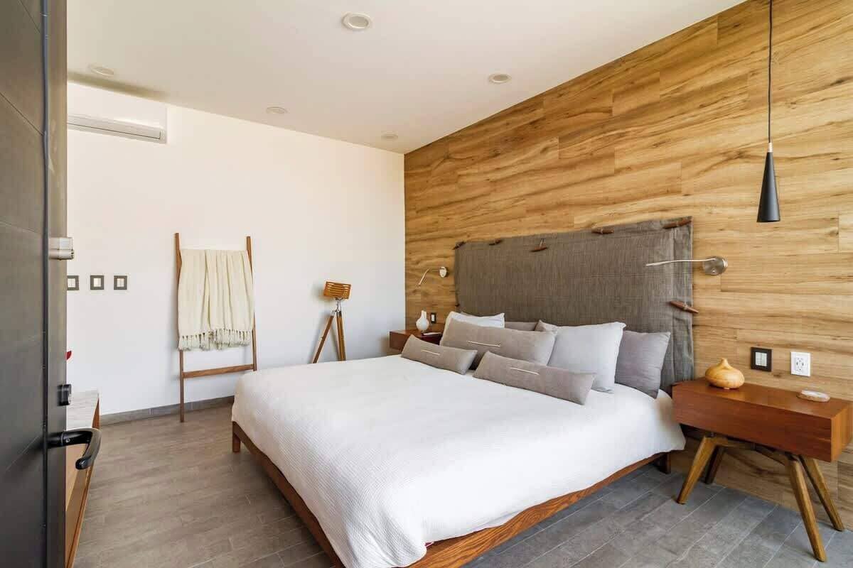 A modern bedroom with a wooden headboard, a large bed with white bedding, a bedside table with a lamp, and a wooden floor.