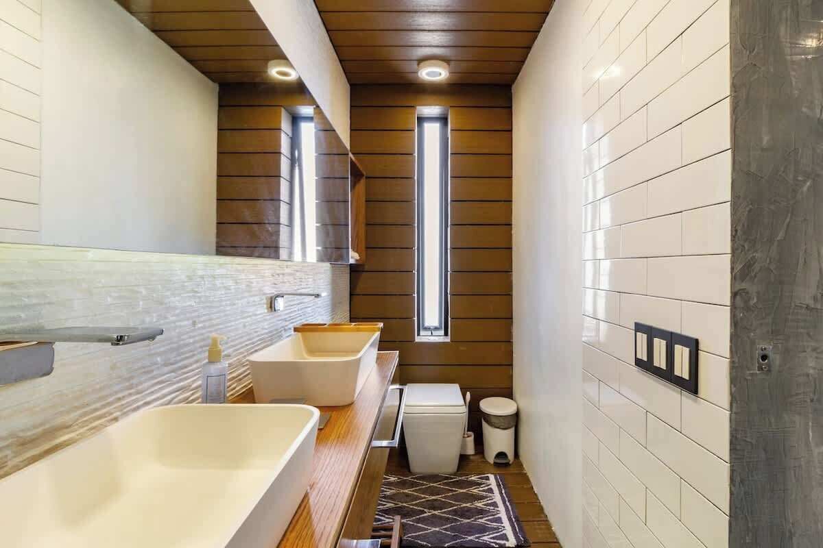 A narrow bathroom with a wooden ceiling, white tiled walls, a large mirror, and a window.
