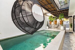 A modern indoor pool with a clear water surface, surrounded by a white wall and a black circular structure. There are potted plants and two white chairs near the pool. - 2 Depas con Alberca Privada Climatizada · Galerías (Zapopan)