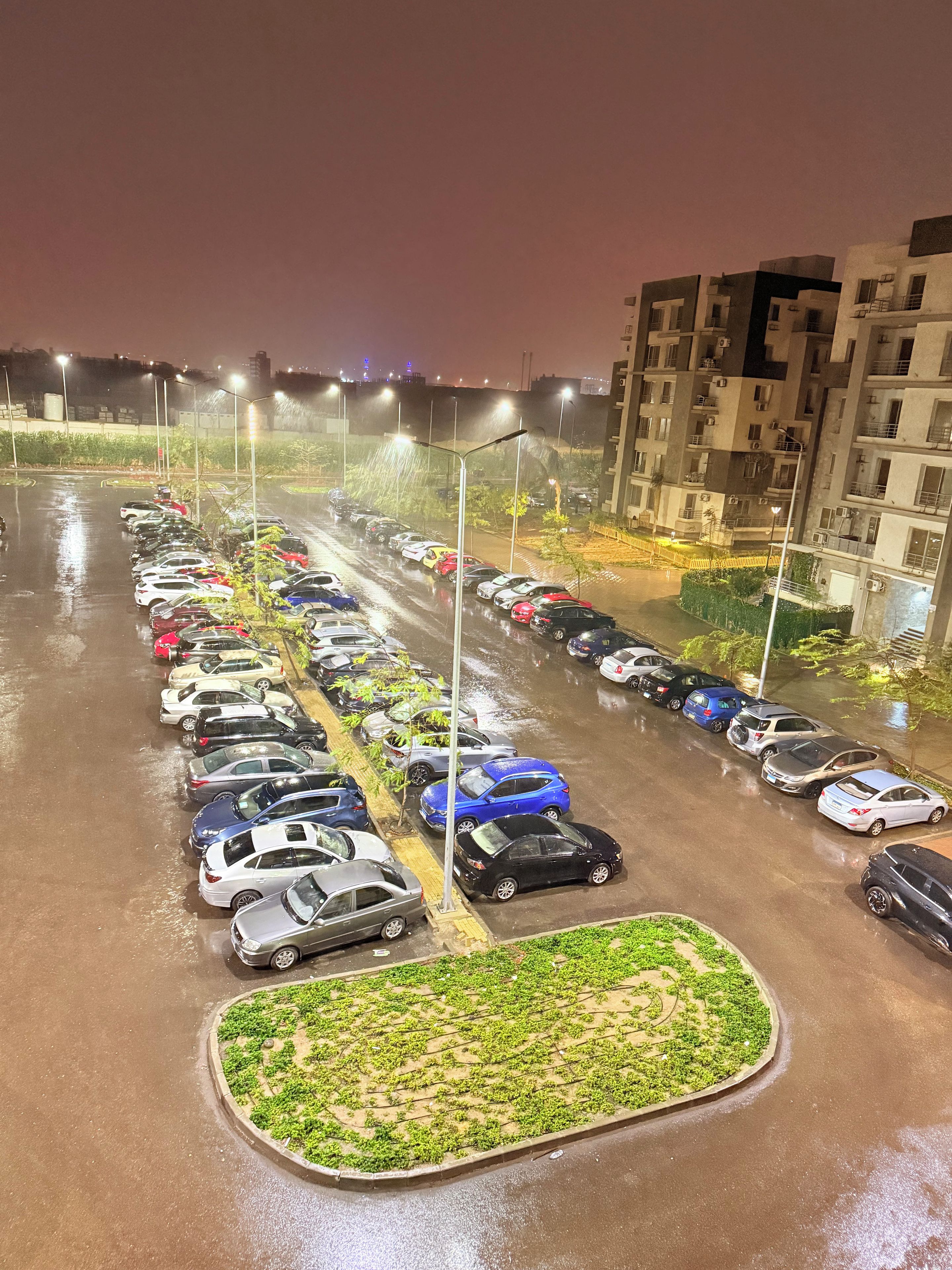 Estacionamento noturno com várias fileiras de carros estacionados e um edifício ao fundo.