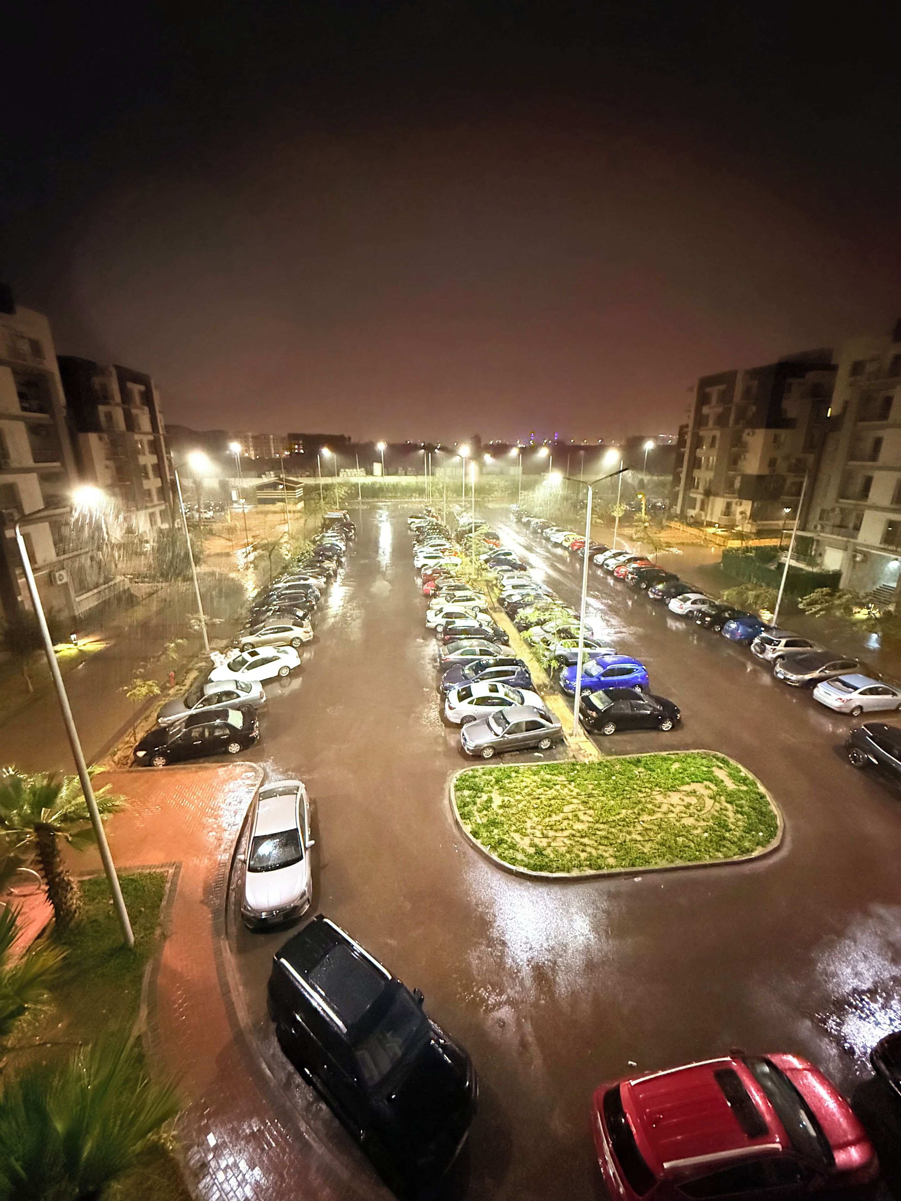 A nighttime parking lot with multiple rows of parked cars and illuminated street lamps.