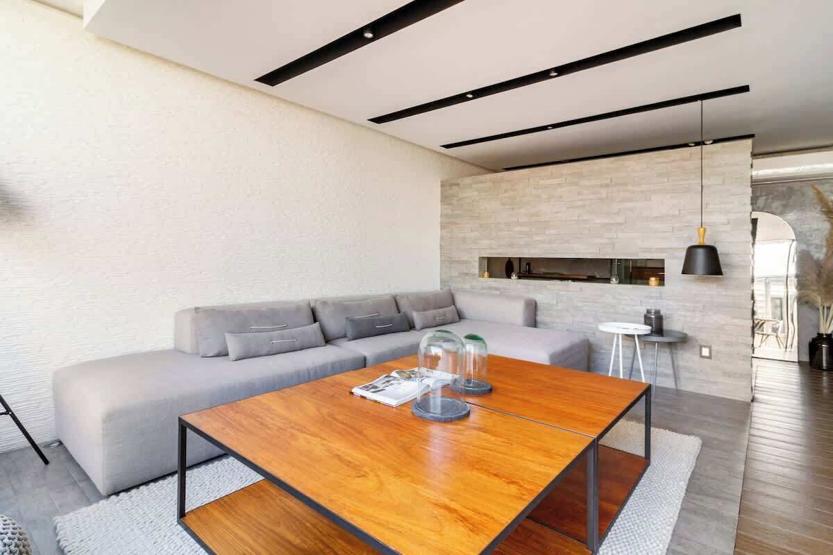 A modern living room with a wooden coffee table, grey sofa, and a stone wall.