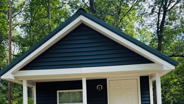 A small, dark blue building with a white door and window, surrounded by trees. - Evergreen Escape 1 bed cabin near Tippy Dam, Trails, Lake Mi, Manistee River NEW (Wellston)