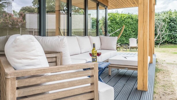 Un espace extérieur moderne, aménagé pour se détendre, avec une terrasse en bois, un canapé avec des coussins et une table sur laquelle se trouvent une bouteille et un verre.